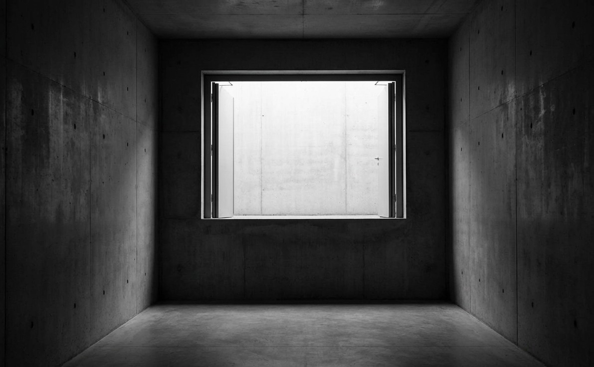 A stark, cinematic, monochromatic black and white photograph of a brutalist concrete hallway. At the end of the hallway is a large, rectangular doorway that is wide open, revealing a bright light. However, the floor of the doorway is 10 feet higher than the hallway floor.